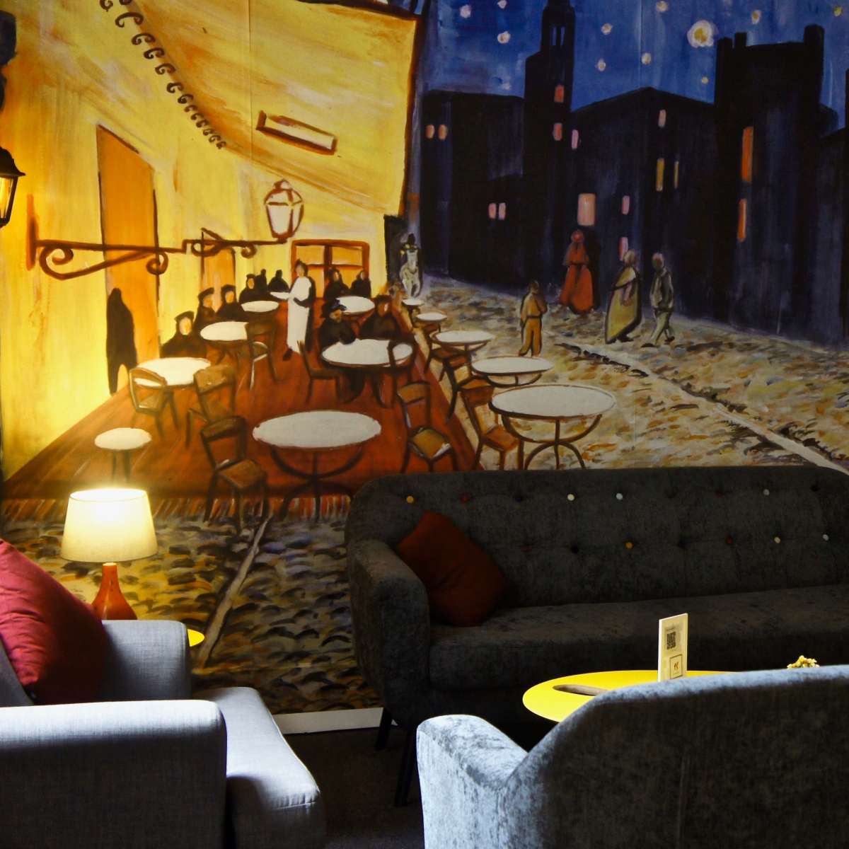 A cozy interior with gray couches placed in front of a vibrant mural depicting a nighttime city scene with people walking along a street lined with café tables.