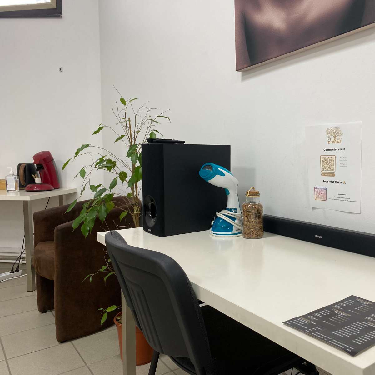 A small workspace features a white table with a black speaker, a spray bottle, a jar of items, and a printed QR code on the wall, alongside a plant and a brown chair.