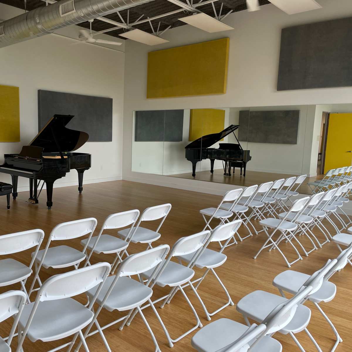 A music rehearsal space featuring two grand pianos, a small table with two chairs, and rows of white folding chairs arranged for an audience.