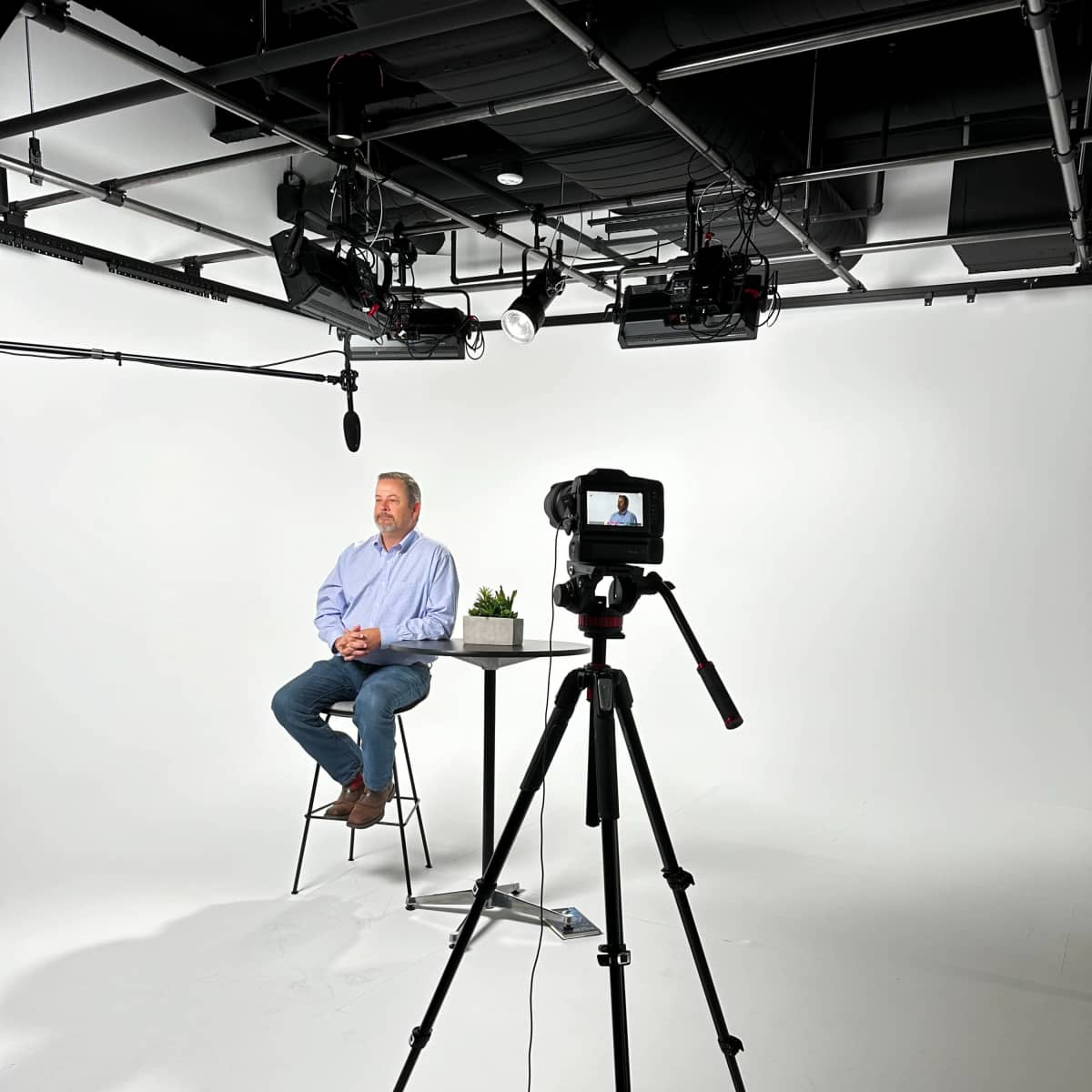 A man sits on a stool in a minimalist studio setting, while a camera on a tripod is focused on him.