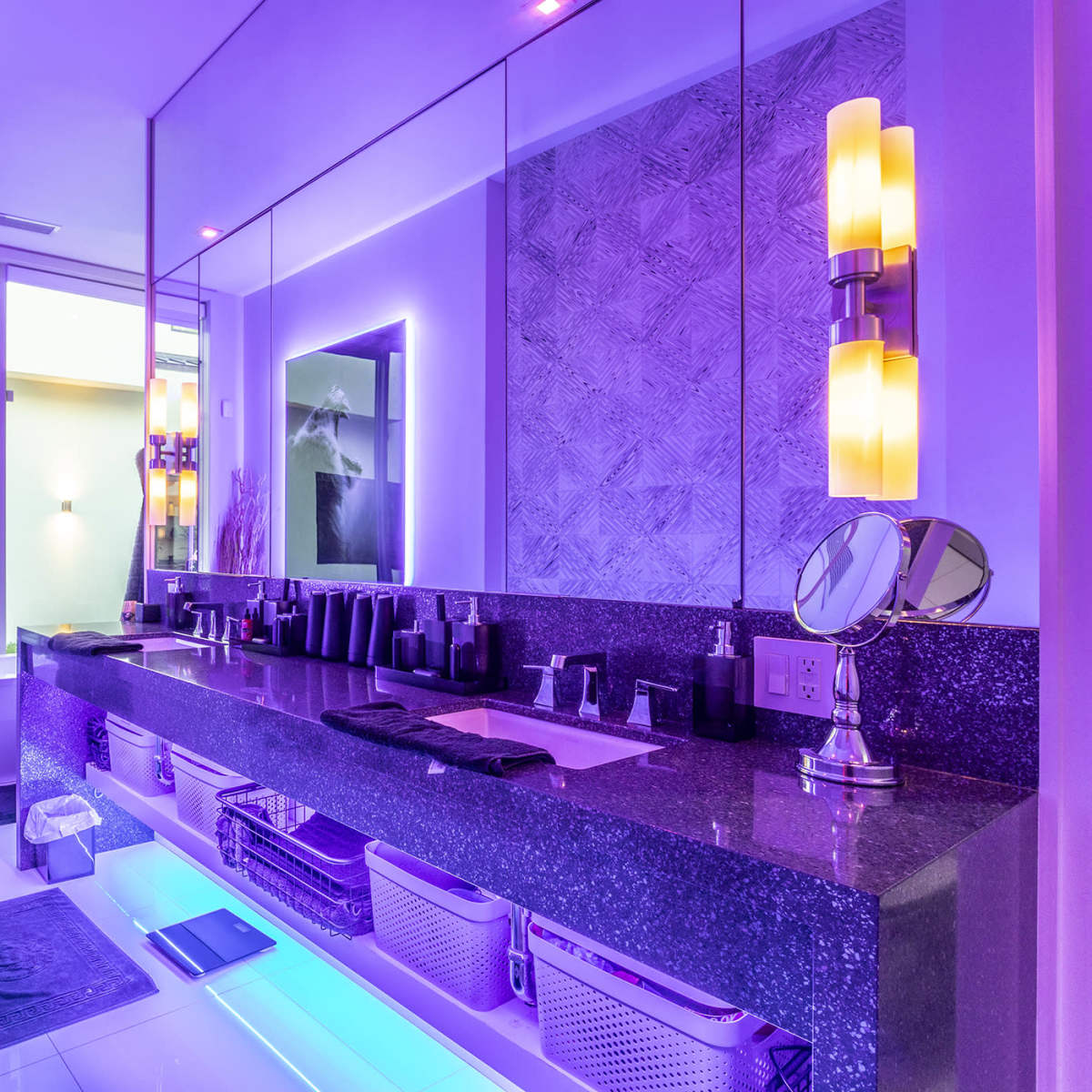 The bathroom features a long countertop with modern fixtures, a plush sofa, and large windows that overlook an outdoor space, all illuminated by purple lighting.