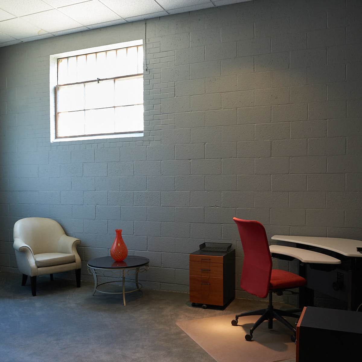 A minimalistic office space features a red chair, a desk, a side table with a decorative vase, and a small filing cabinet against a gray wall.