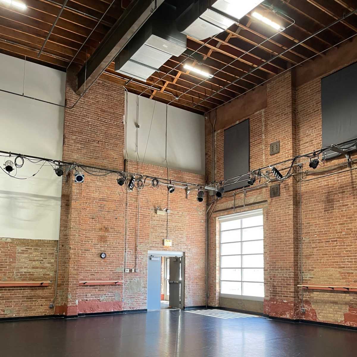 A spacious dance studio with brick walls, wooden beams, and a large door leading outside.
