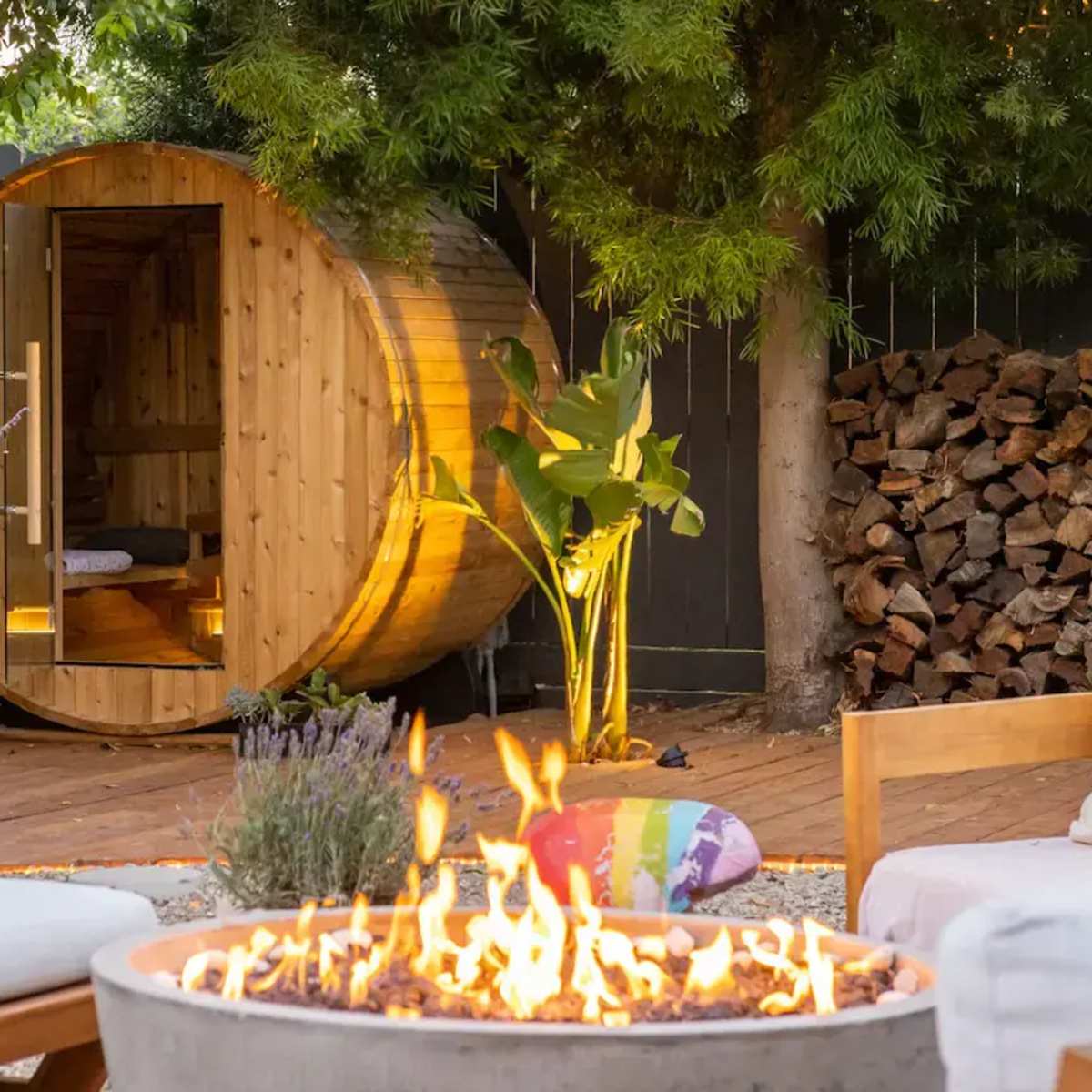 A wooden barrel sauna sits in a backyard surrounded by plants, with a fire pit in the foreground and a stacked woodpile in the background.