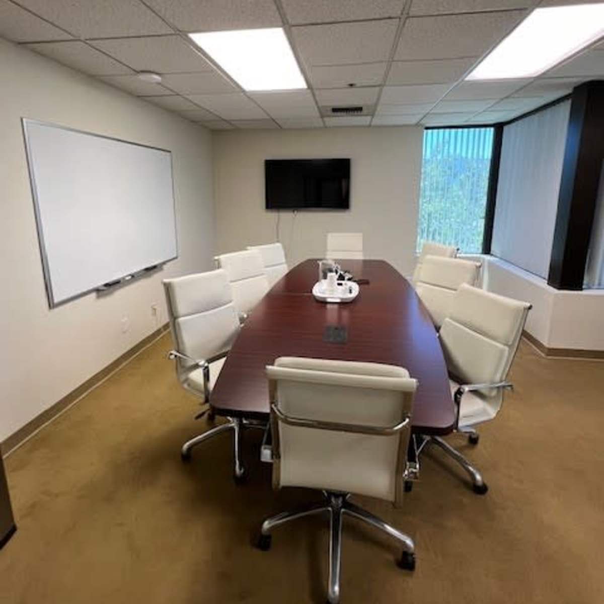 A conference room with a large wooden table surrounded by eight white leather chairs, a whiteboard, and a wall-mounted TV.