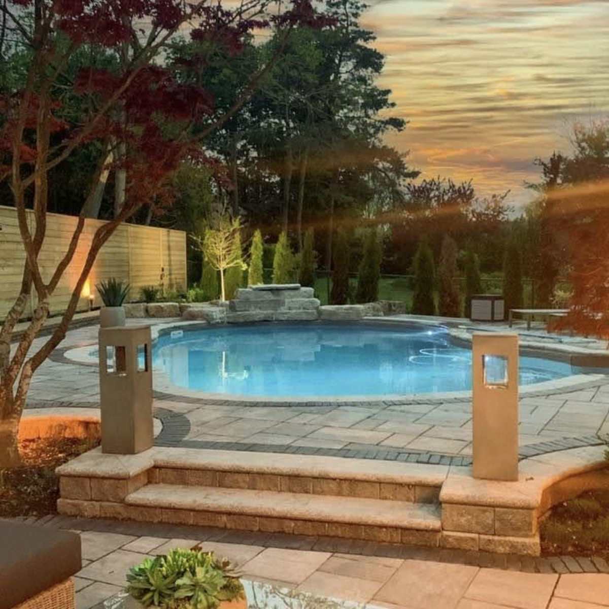 A landscaped backyard with a circular swimming pool, surrounded by stone pathways and lit by modern outdoor lanterns, under a sunset sky.