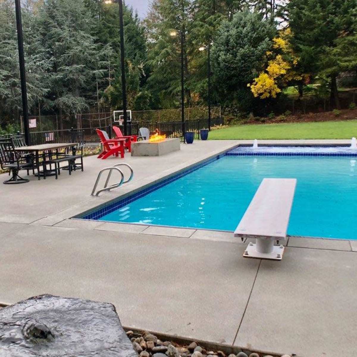 A swimming pool surrounded by a patio area with a diving board, lounge chairs, and trees in the background.