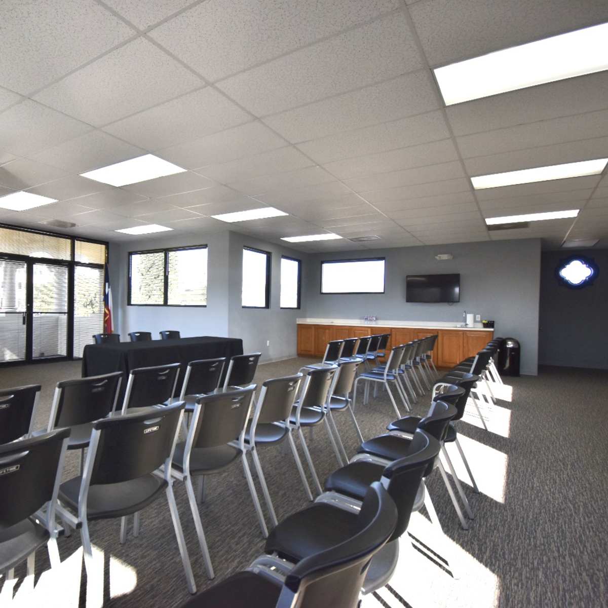 A spacious meeting room with rows of black chairs facing a presentation area, equipped with windows and a television screen.