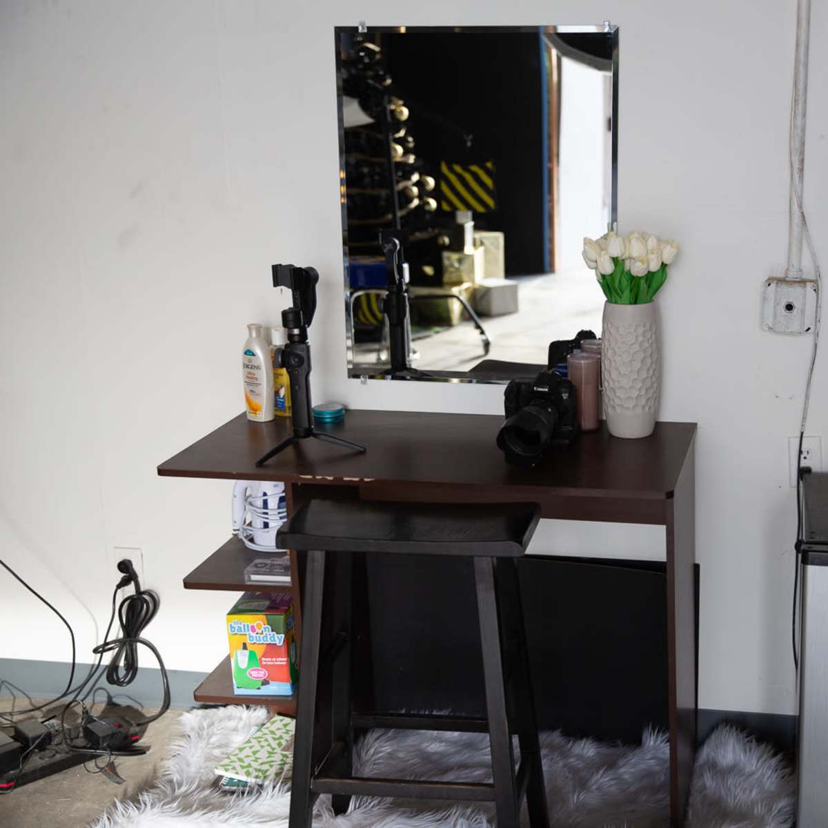A small wooden vanity with a mirror and a vase of white flowers is positioned against a wall, next to a mini refrigerator and a stack of boxes.