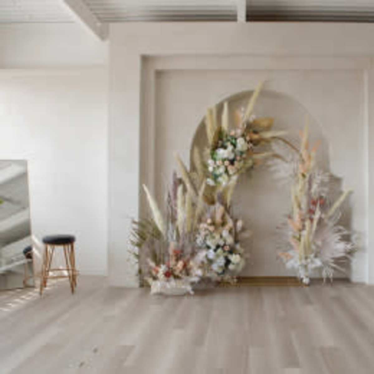 A minimalist interior space with a large arch of decorative dried flowers and plants against a light-colored wall, accompanied by a mirror and a stool.