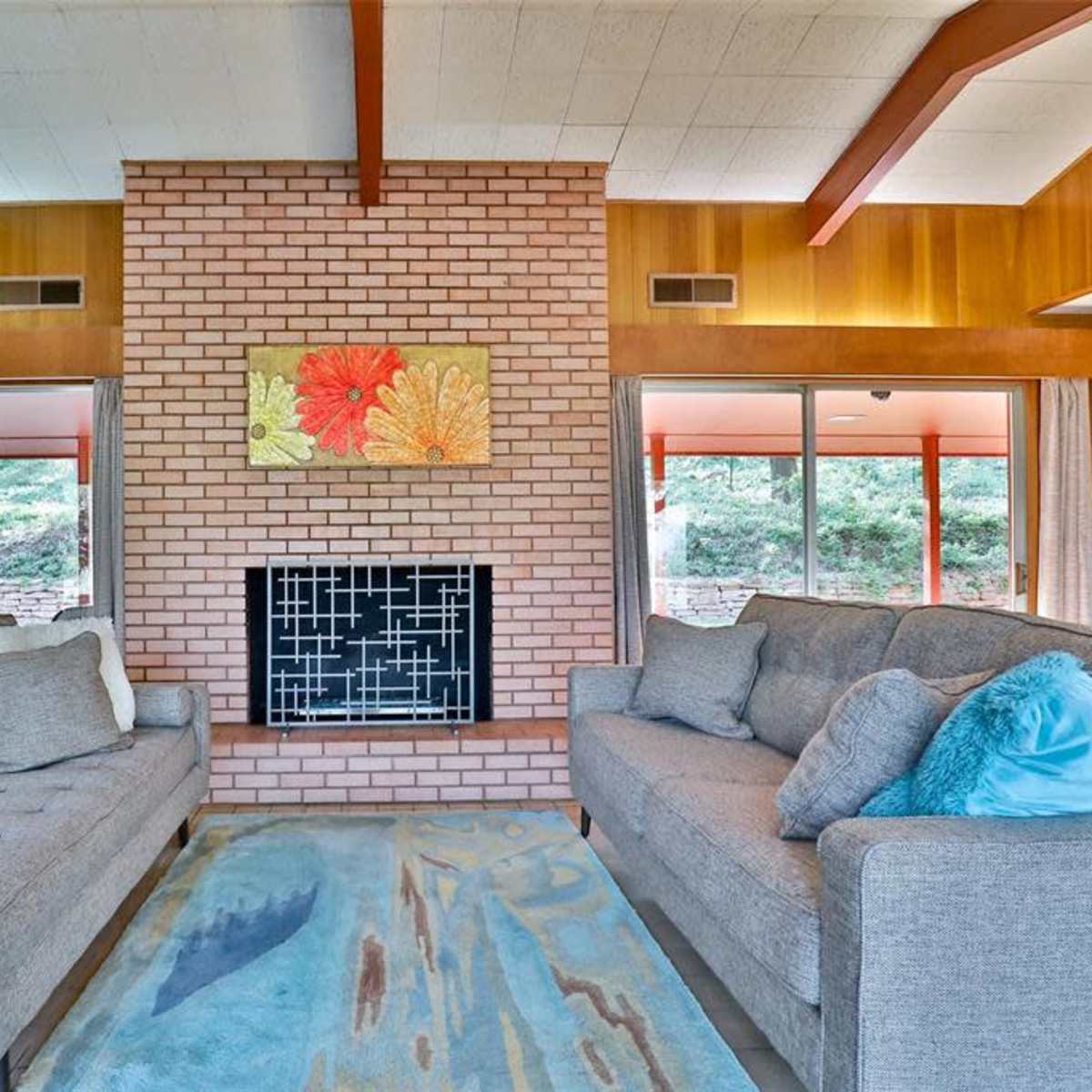 The room features two gray sofas set opposite a brick fireplace, with large windows that provide a view of the outdoors.