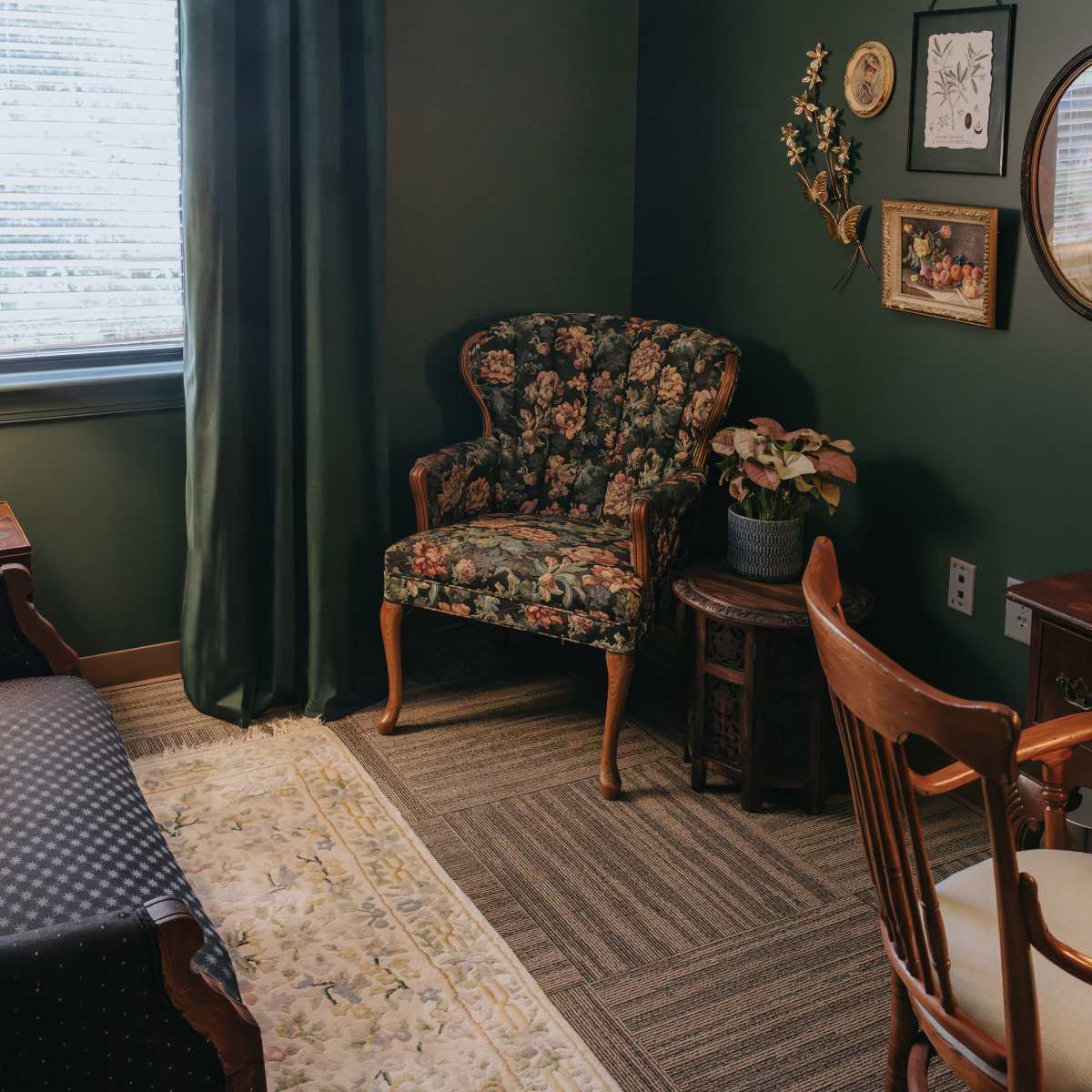 A cozy room with a floral armchair, a small wooden table, a mirror, and a vintage-style lamp beside a sofa on a patterned rug.