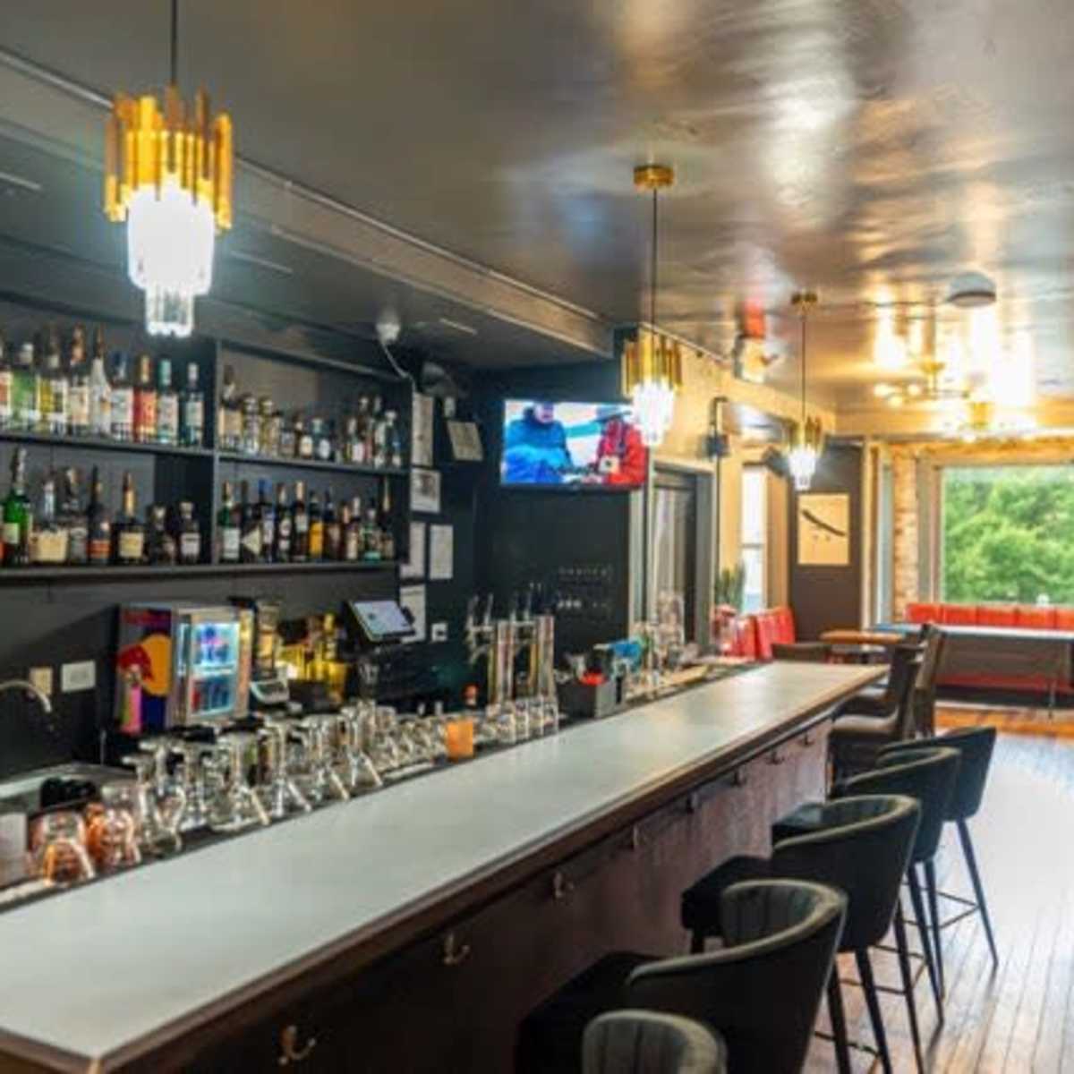 A modern bar interior with a light-colored countertop, a variety of bottles on shelves, and several bar stools lined up along the counter.