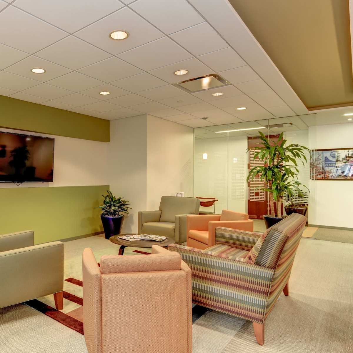 A waiting area features two green armchairs and two orange armchairs arranged around a coffee table, with a television mounted on the wall and plants in the corners.