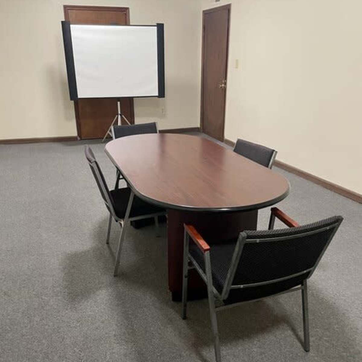 A rectangular table with four chairs is positioned in a sparsely furnished meeting room with a projection screen at one end.