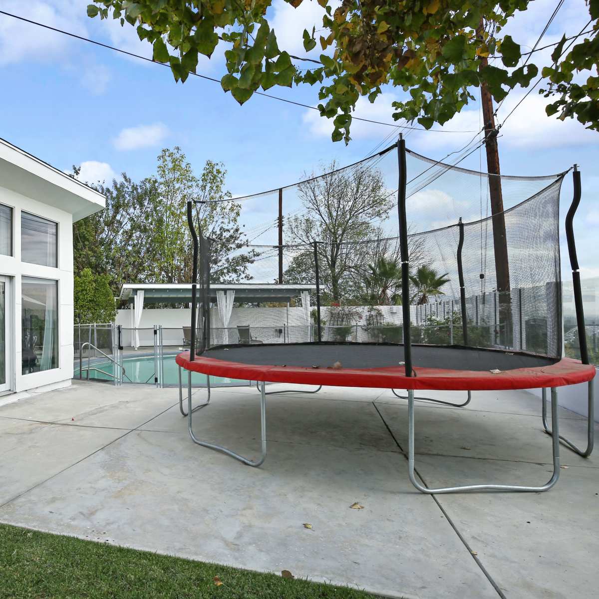 A modern house with large windows adjacent to a trampoline and a swimming pool surrounded by concrete and greenery.