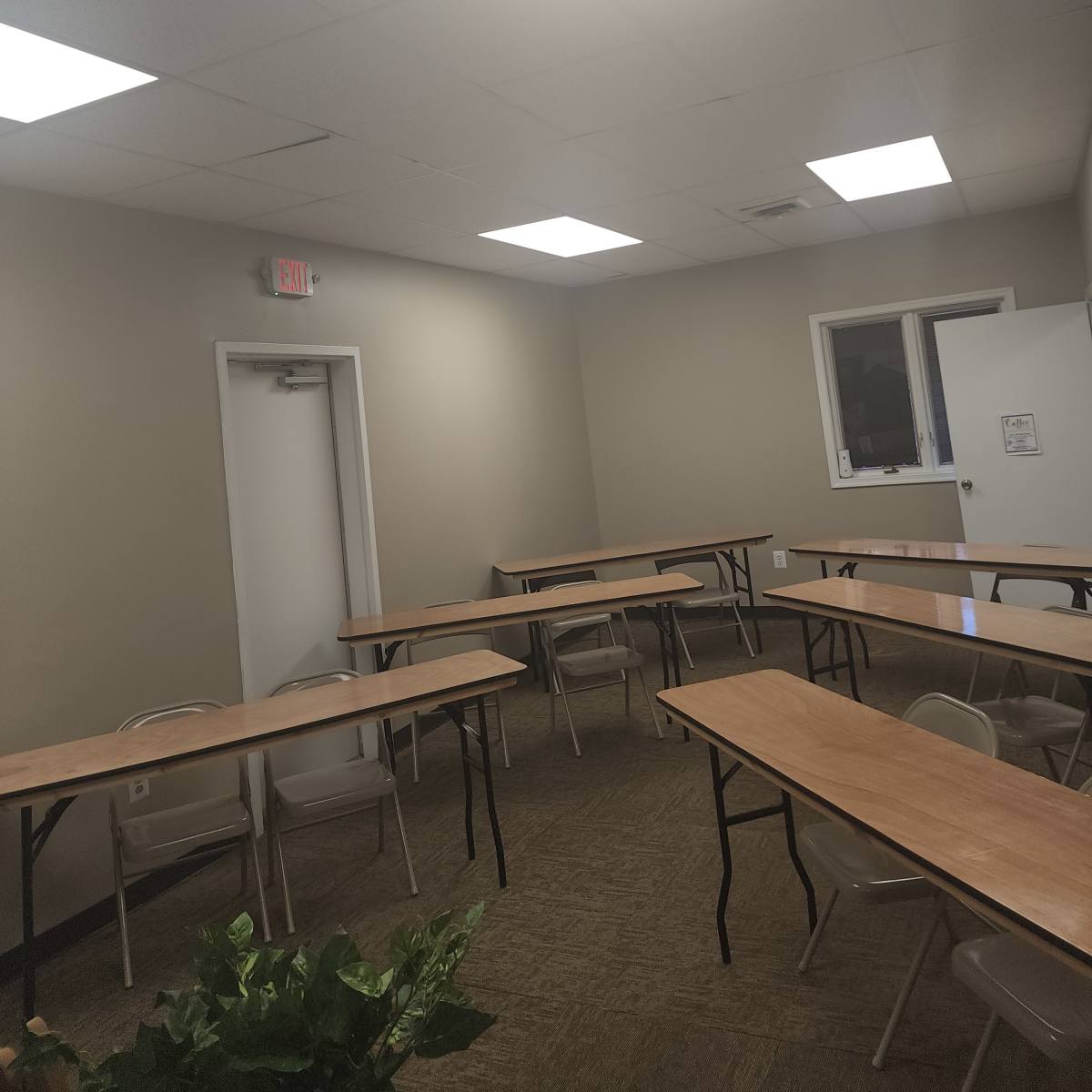 A small meeting room with several tables arranged in a U-shape, beige walls, and bright fluorescent lighting.