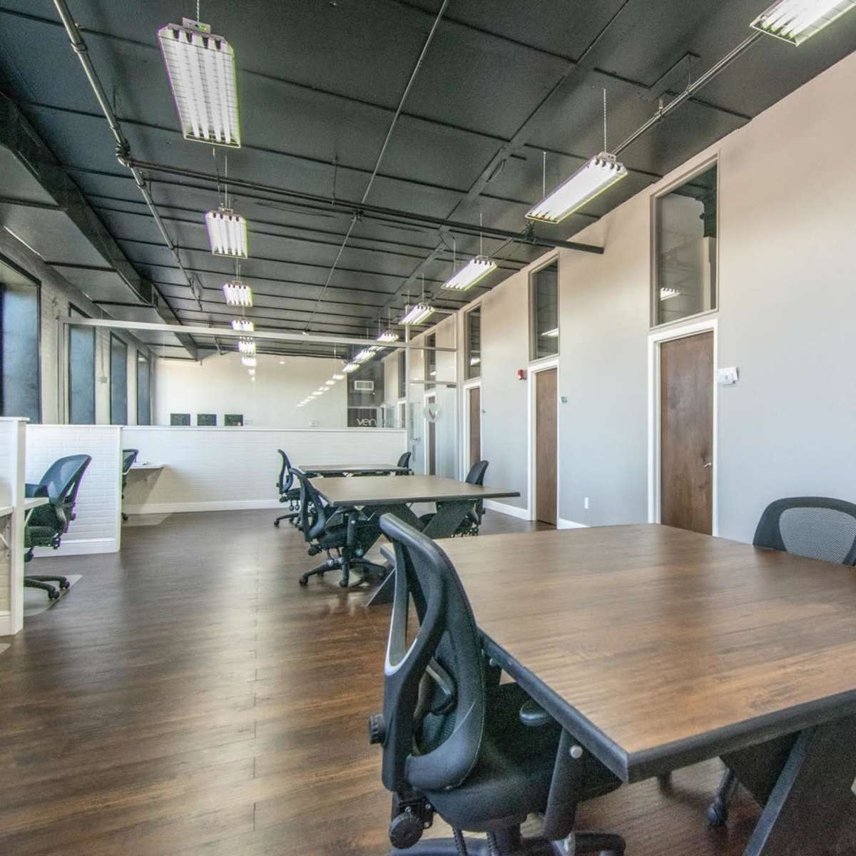An empty office space with multiple wooden tables and black office chairs, featuring large windows along the walls.