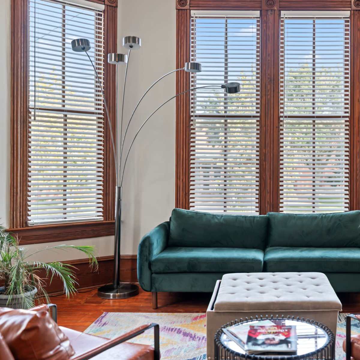 A green sofa sits in a bright room with large windows and wooden blinds, accompanied by a light-colored ottoman and two brown armchairs.