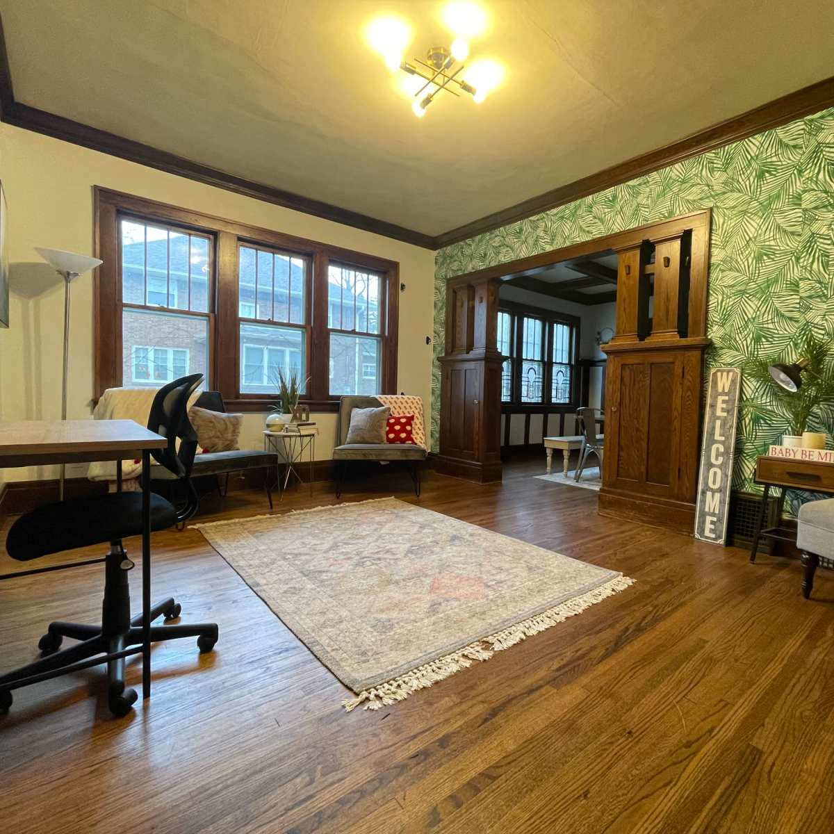 A well-lit living room with a patterned rug, a desk and chair, seating areas, and a green leafy wallpaper.