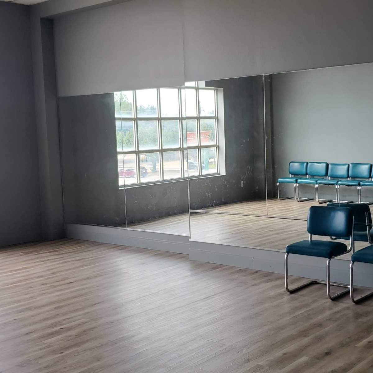 An empty room with wooden floors, a large mirrored wall, and a row of blue chairs arranged against the opposite wall.