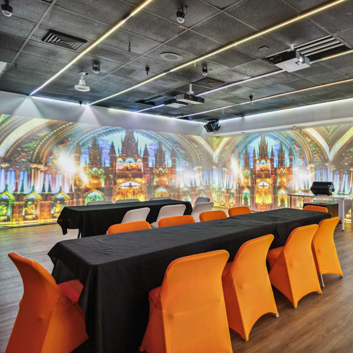 A conference room features a long table with black covers and bright orange chairs, illuminated by colorful projections of a cathedral interior on the walls.
