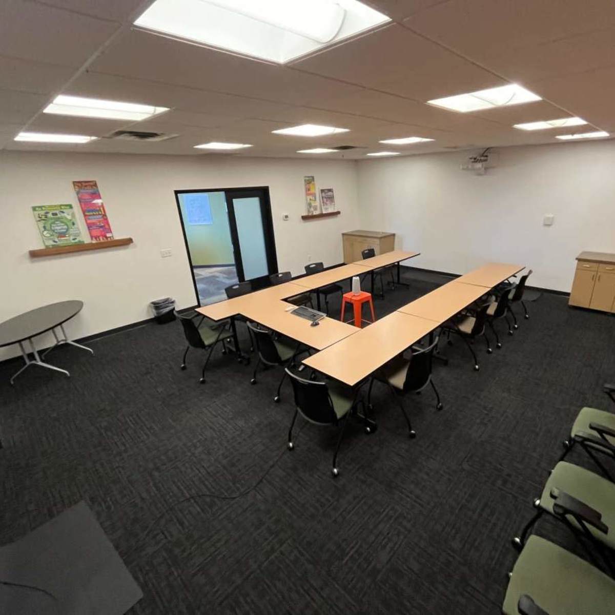 A spacious meeting room with several tables arranged in a U-shape, surrounded by chairs and a cabinet against one wall.