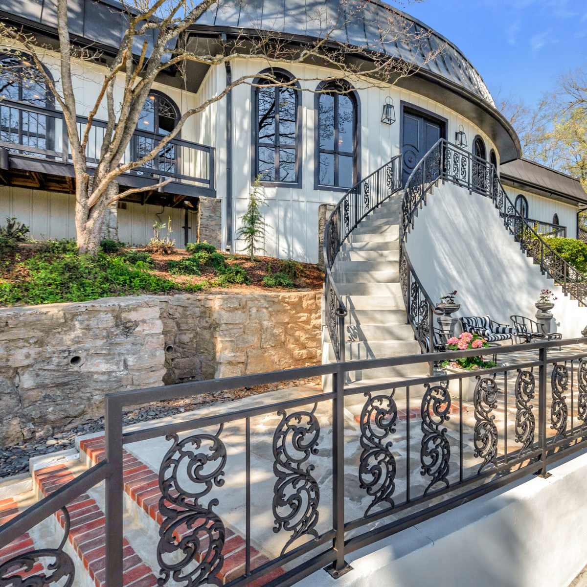 A modern house with a circular design, featuring a stone staircase leading up to an ornate black railing and landscaped surroundings.