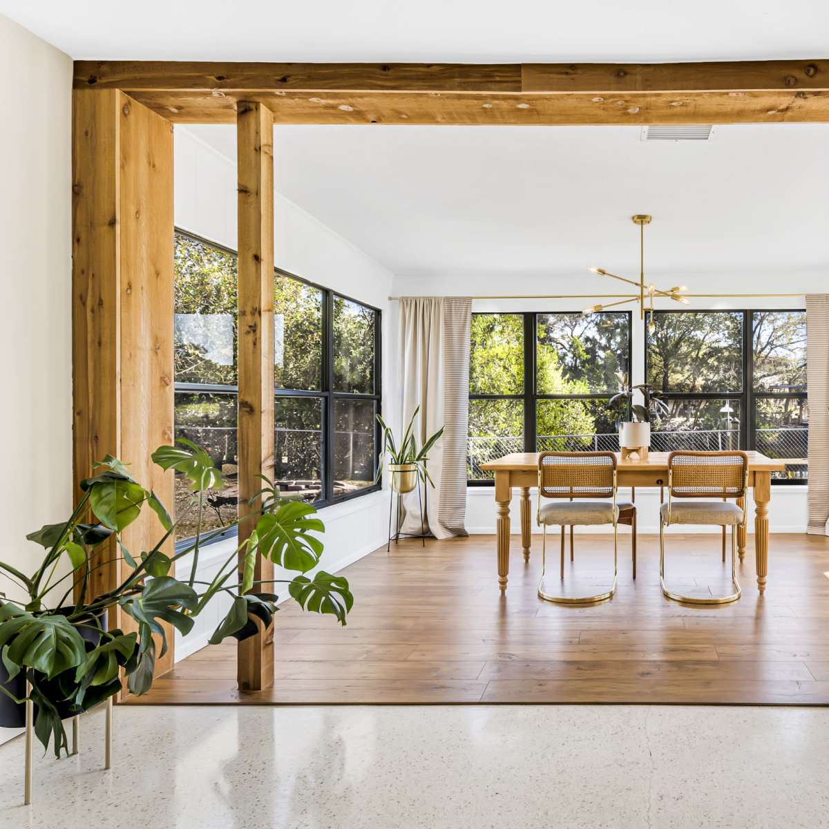 A spacious dining area features a wooden table surrounded by chairs, framed by large windows and a view of greenery outside.