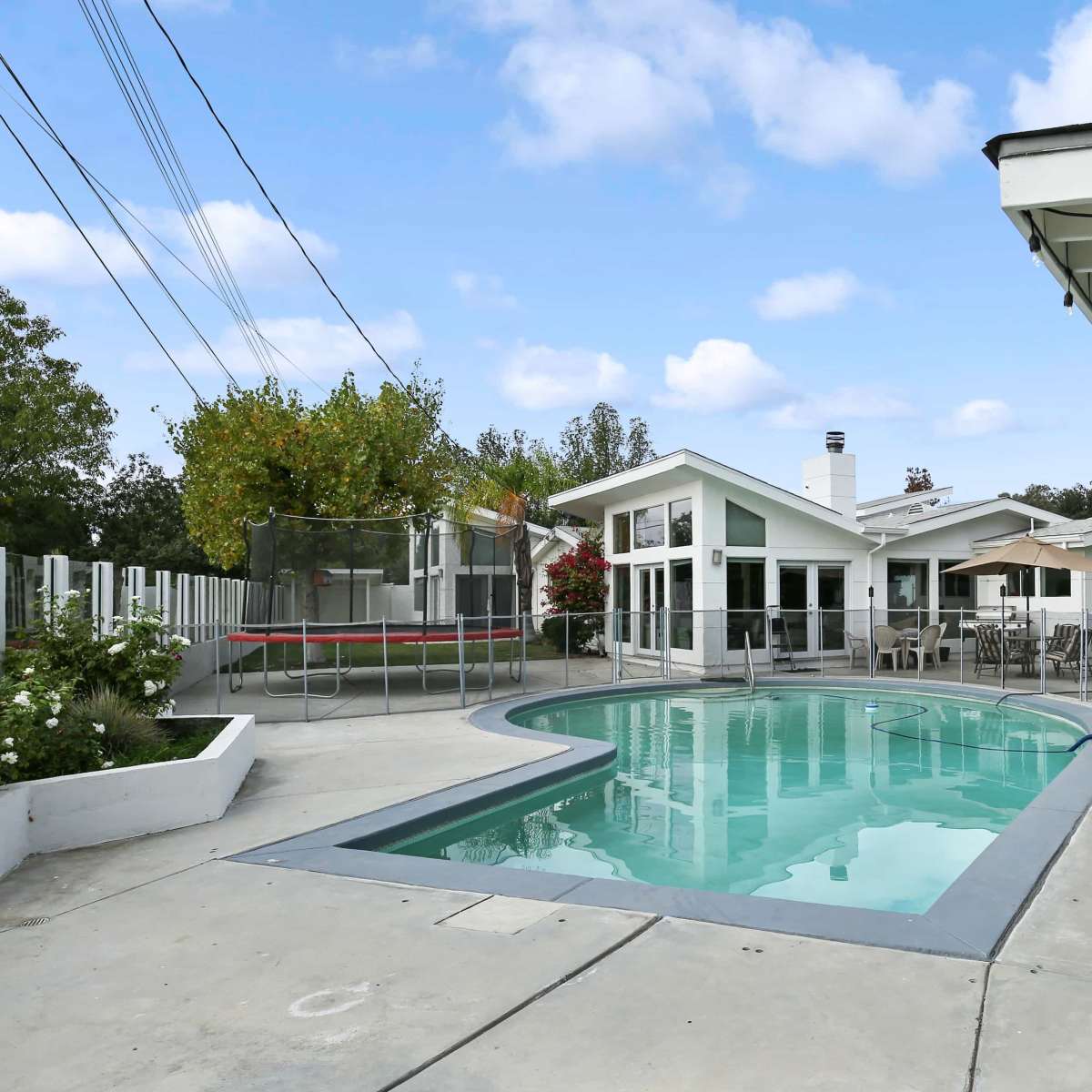 A backyard with a swimming pool, surrounded by a patio area and a trampoline, alongside a modern-style house.