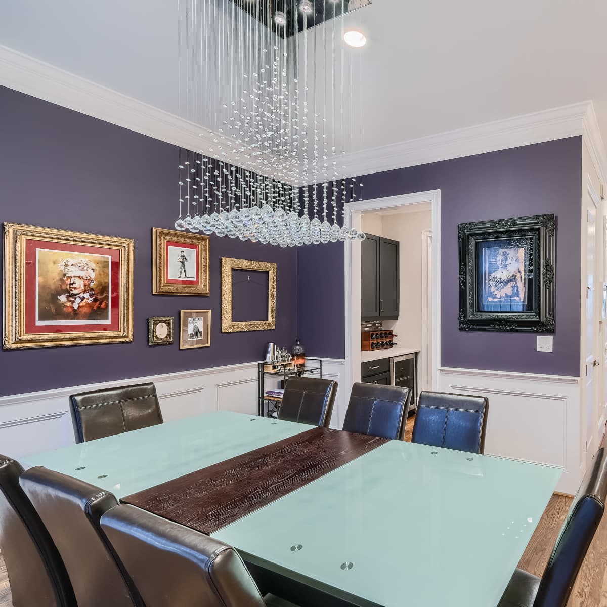 A modern dining room with a glass-topped table surrounded by black chairs, framed artwork on the purple walls, and a staircase leading to another level.