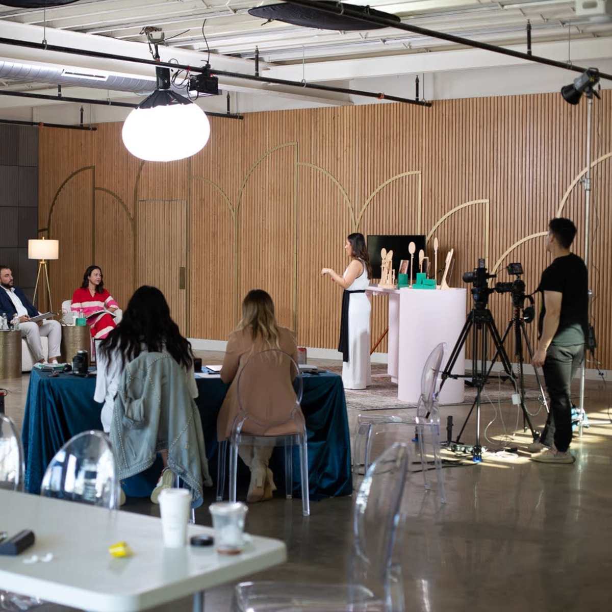 A group of people are seated in a studio, observing a presenter who is speaking in front of a wooden backdrop with sculptures, while cameras are set up to capture the event.