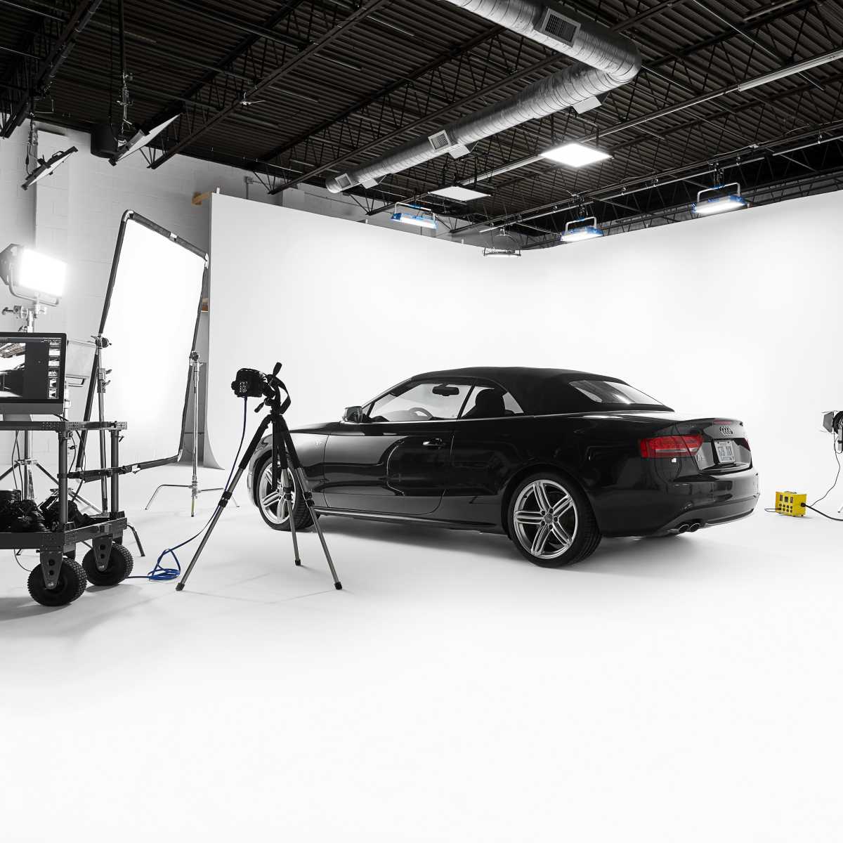 A black convertible car is positioned on a white backdrop in a photography studio equipped with lights and a camera setup.