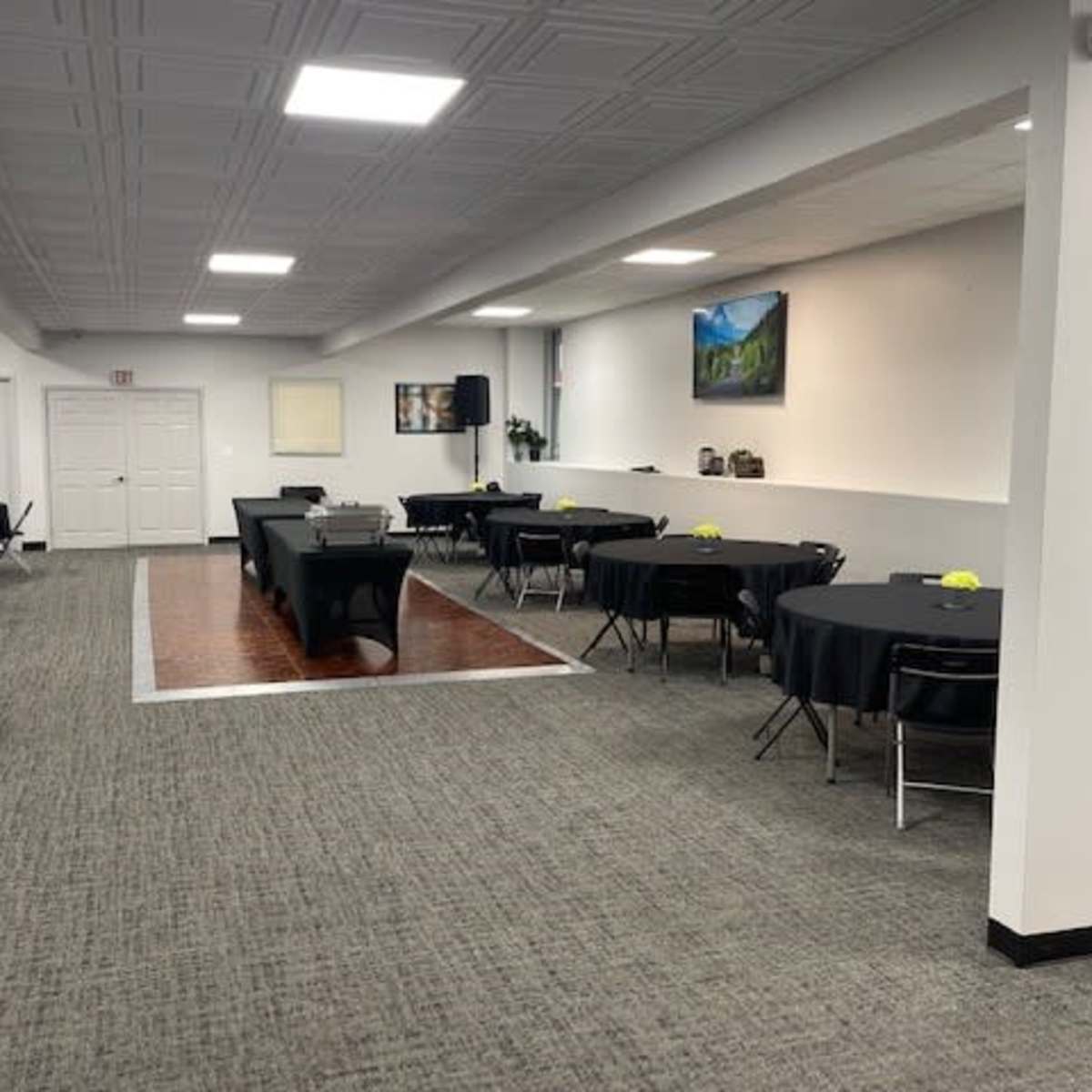 A spacious event room with several round tables set up around the perimeter and a central rectangular table, illuminated by ceiling lights.