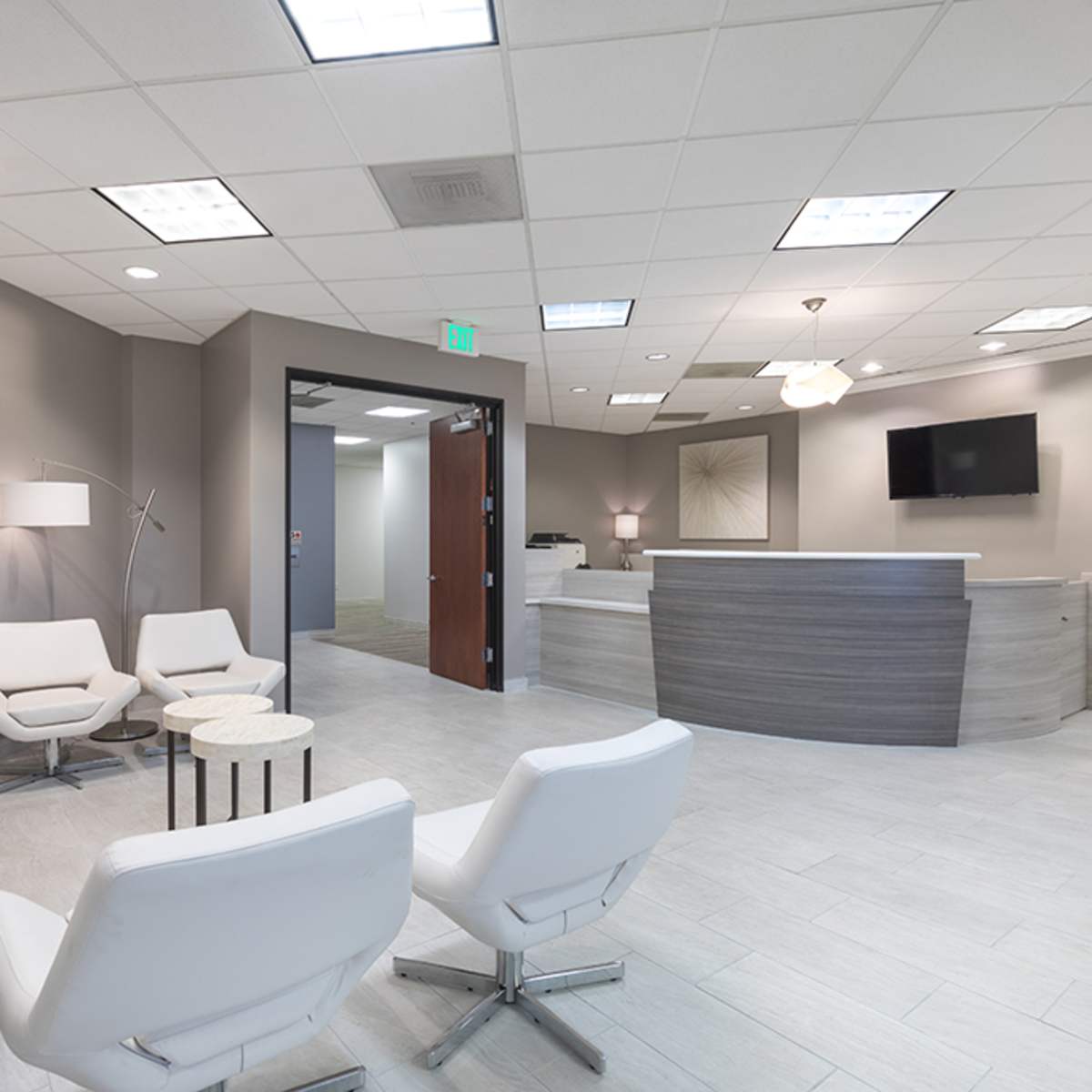 A modern waiting room features several white chairs and small tables, with a reception desk in the background and a flat-screen TV mounted on the wall.