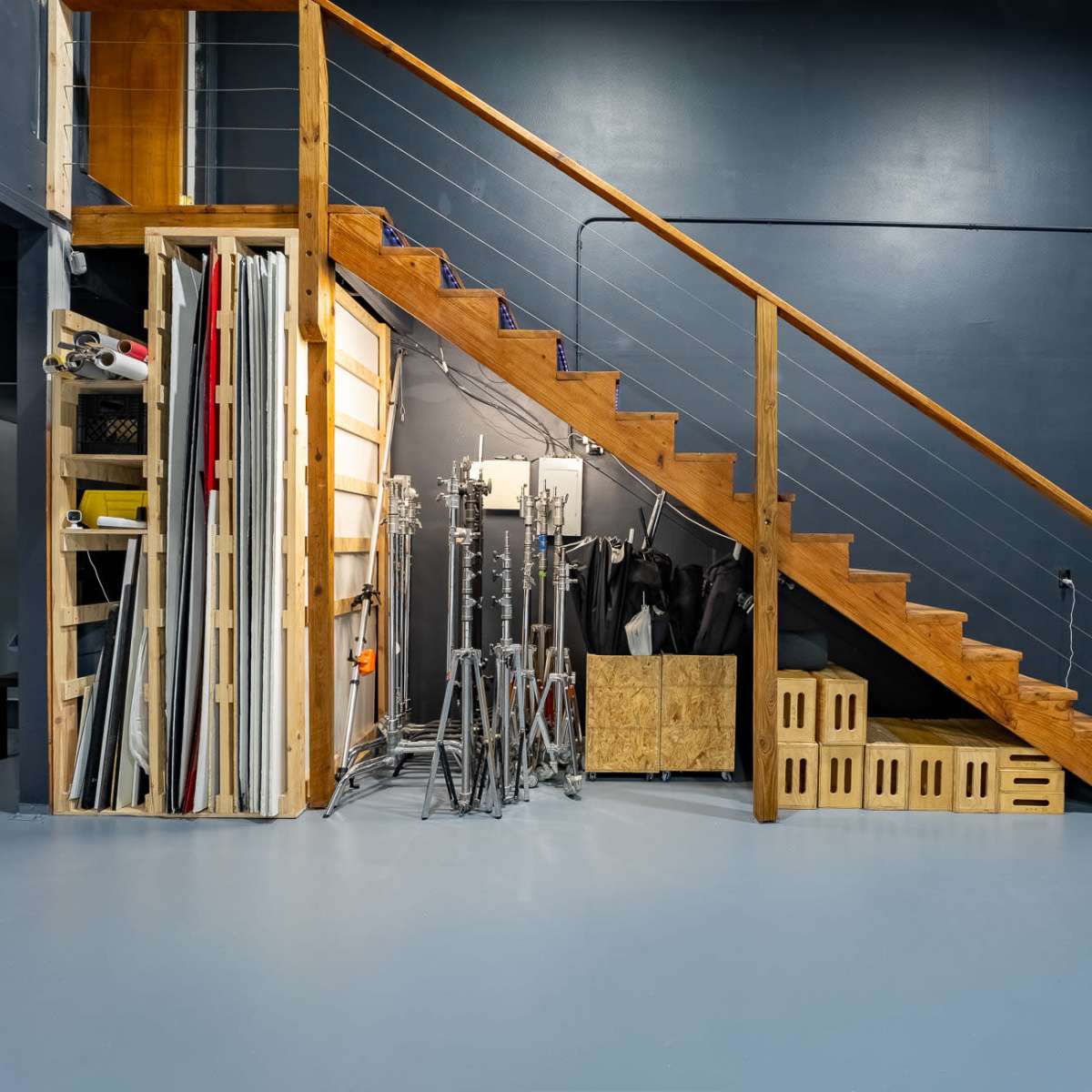 A studio space with wooden stairs leading to a higher level, alongside organized photography equipment and rolls of backdrop materials.