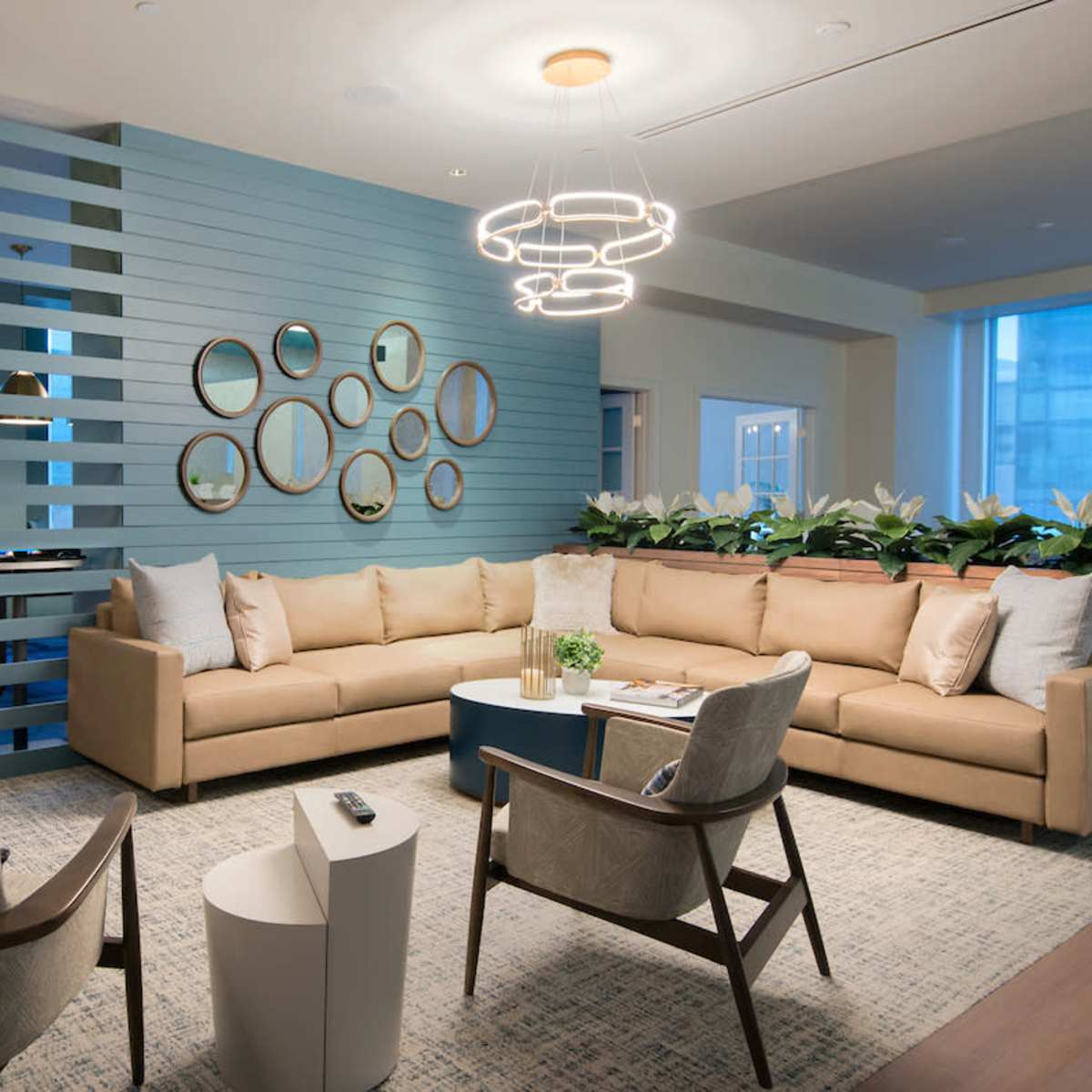 A modern lounge area features a beige sectional sofa, circular mirrors on the wall, and a round coffee table, illuminated by a contemporary light fixture.
