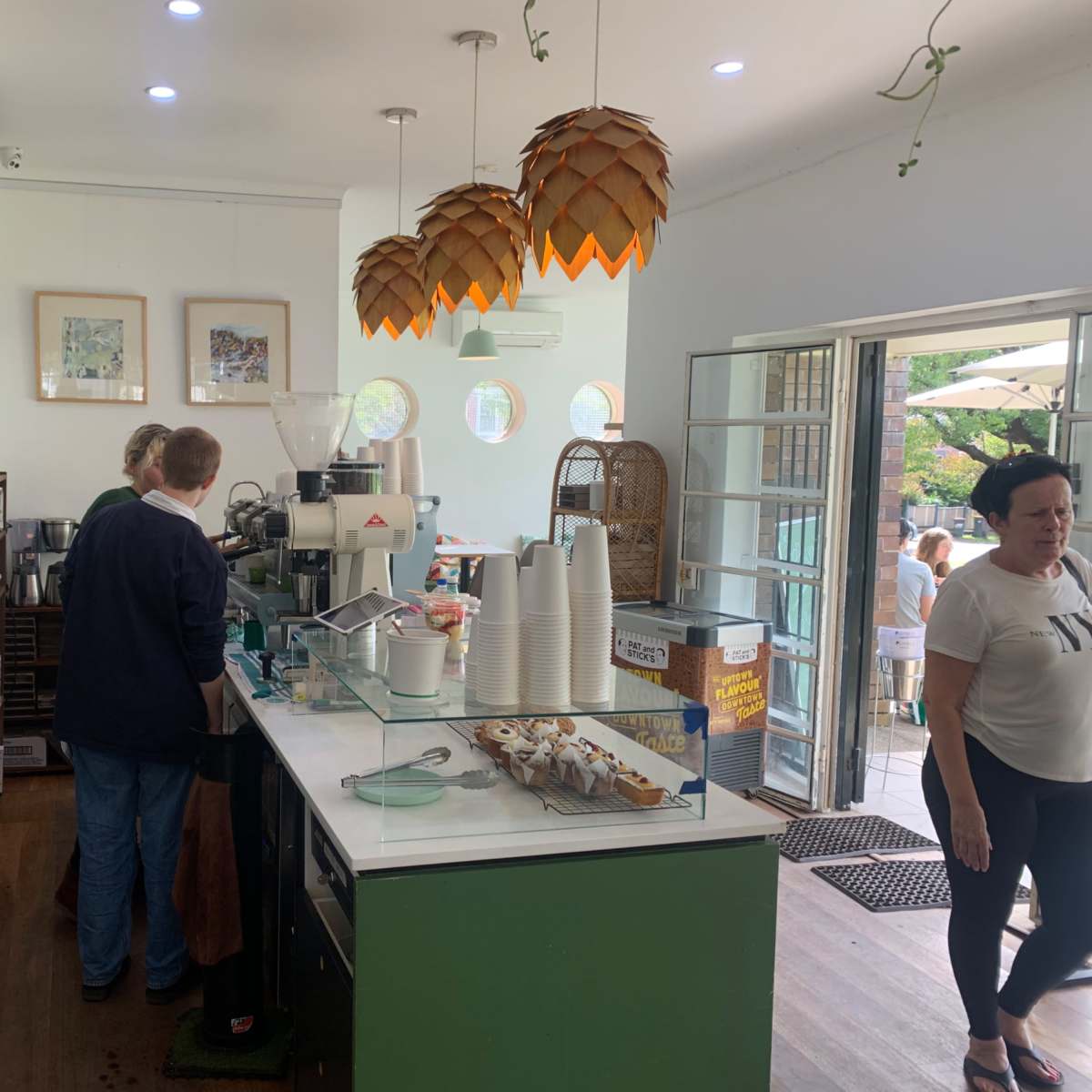 A coffee shop interior features two baristas behind the counter, surrounded by various coffee-making equipment and display items, with patrons visible outside through an open door.