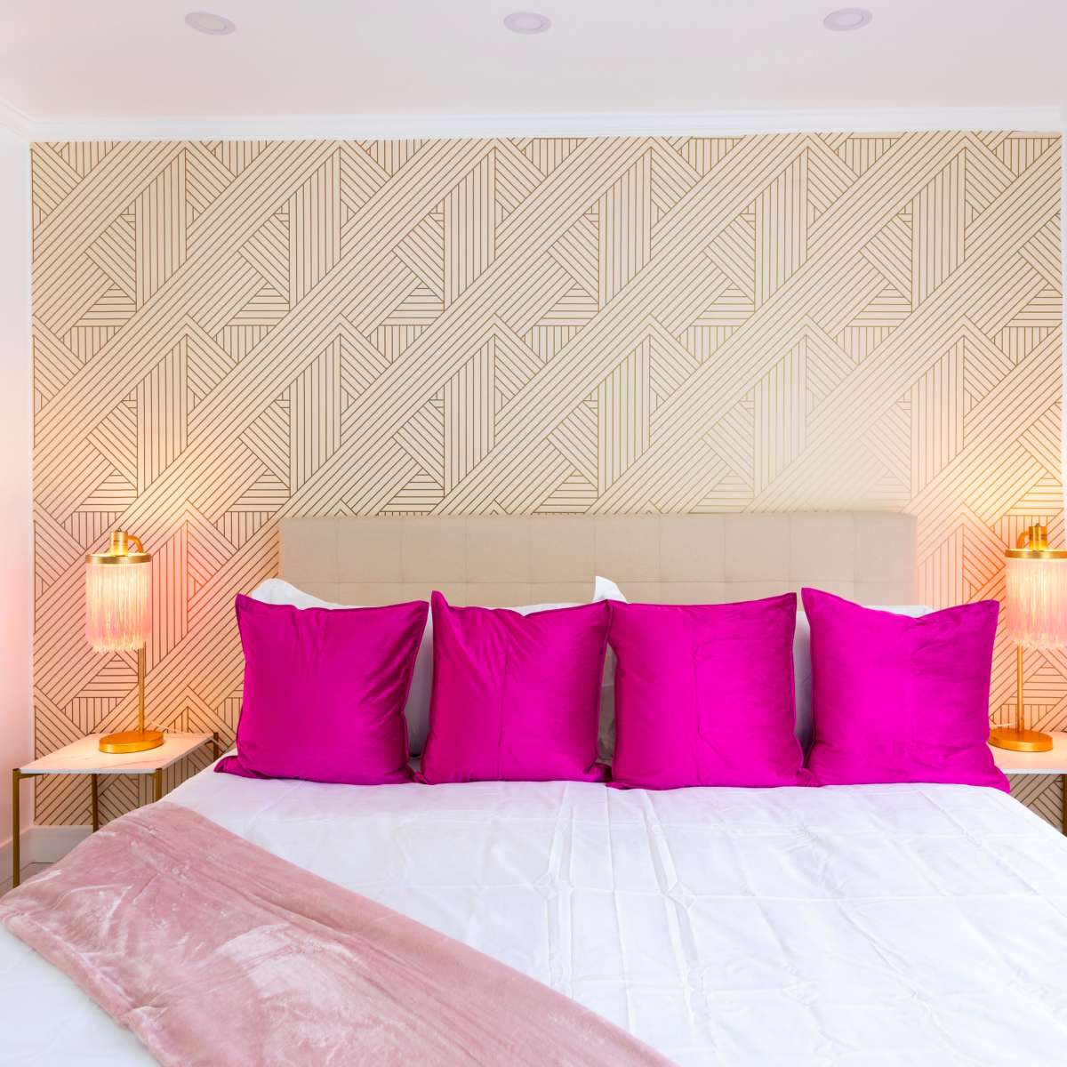 A neatly arranged bedroom featuring a bed with a beige headboard, adorned with five pink pillows, against a patterned wall, with two bedside lamps on either side.