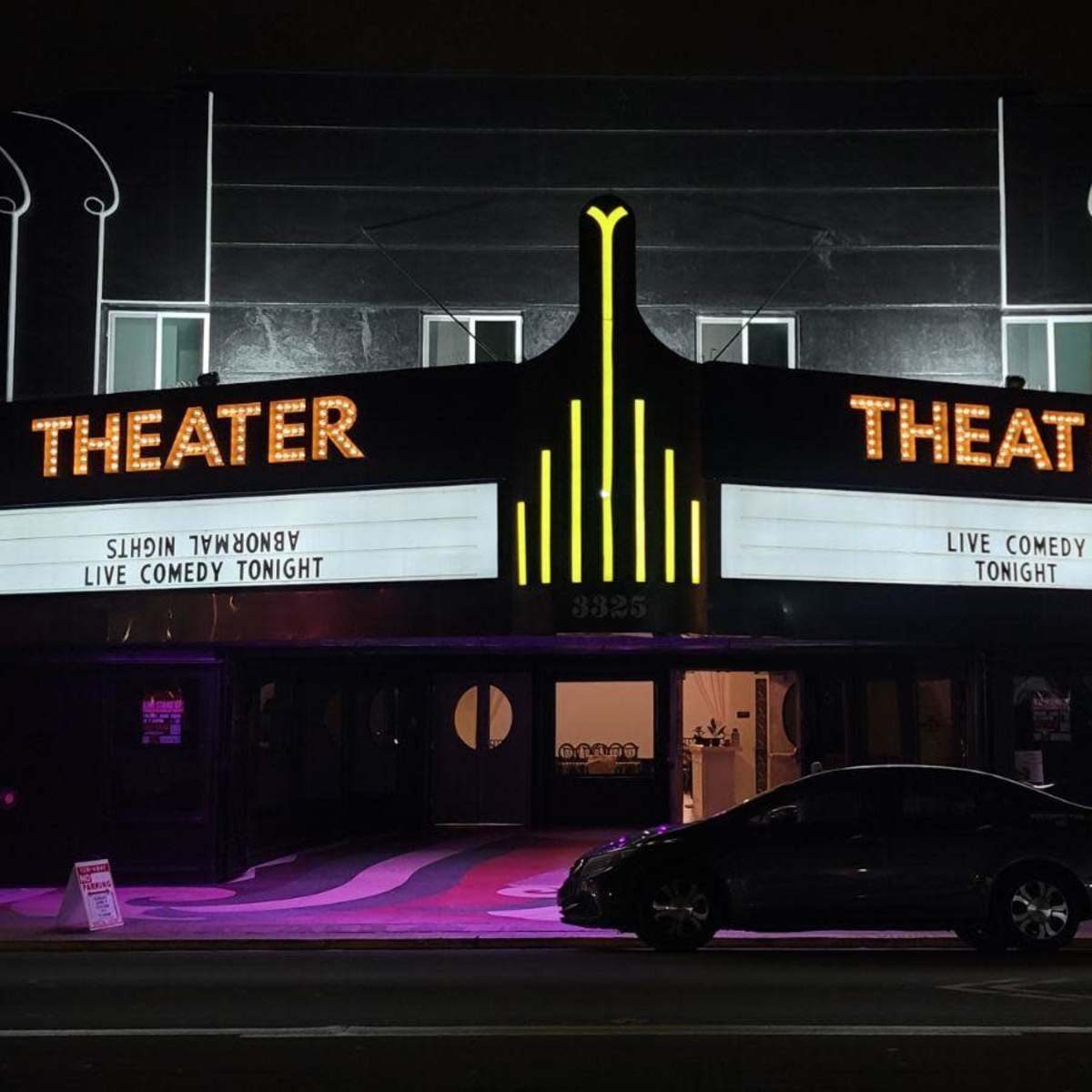 A theater building displays a marquee announcing a live comedy event, with a sleek, illuminated facade and a parked car in front.