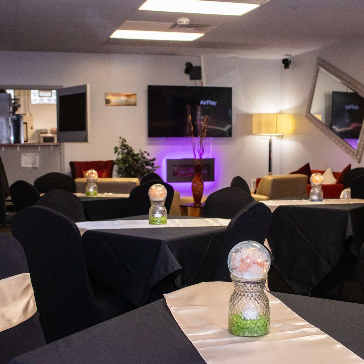 A restaurant interior with dark tables covered in black cloths and decorative centerpieces, featuring a TV, a lamp, and mirrors on the walls.