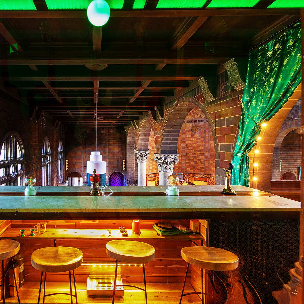 A vibrant, multi-level bar area featuring a wooden counter, high stools, and decorative lighting against a backdrop of brick arches and lush drapes.