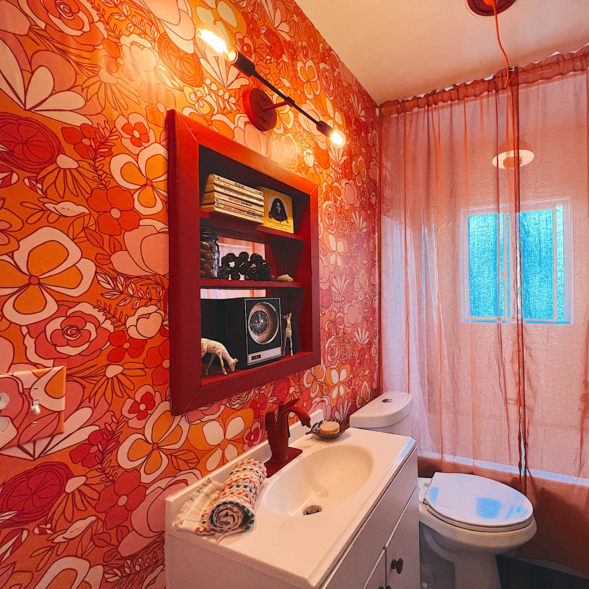 A small bathroom with floral-patterned wallpaper, a white vanity with a sink, a toilet, and a window covered by a sheer curtain.