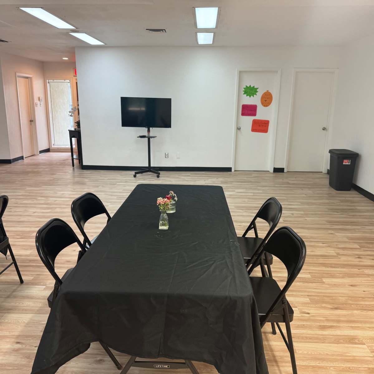 A spacious room with a central black table set for a gathering, surrounded by chairs, and featuring a television and brightly colored posters on the walls.