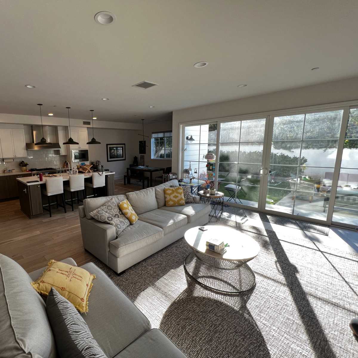 A modern living area features a light-colored sofa, a round coffee table, and an open kitchen with bar seating, all illuminated by natural light streaming through large windows.