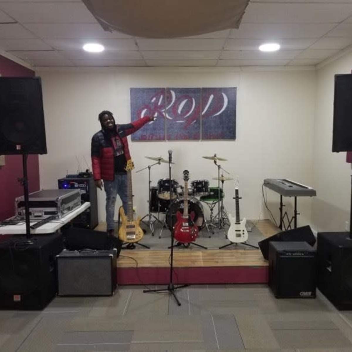 A man stands on a wooden stage in a music rehearsal space surrounded by musical instruments and audio equipment.