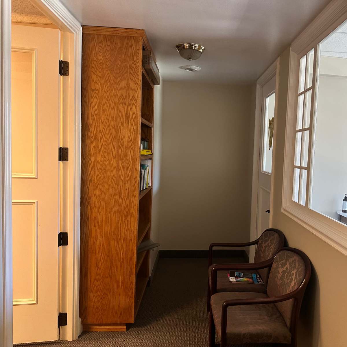 A hallway with light-colored walls, a wooden bookcase on the left, and two chairs positioned along the side, leading to a door at the end.