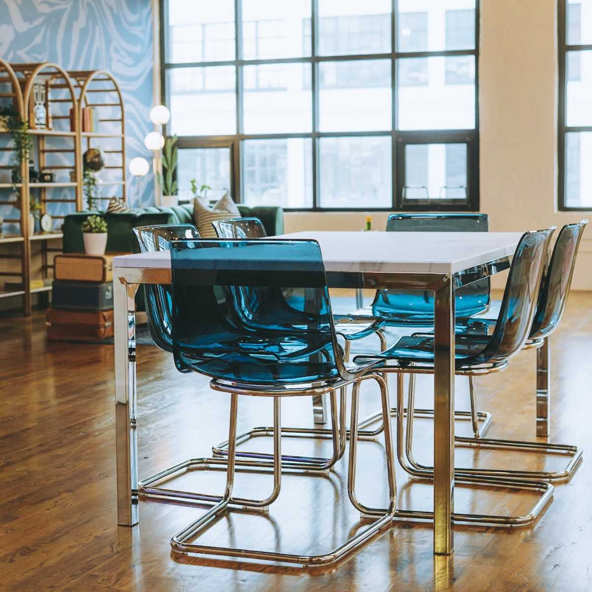 A modern dining area features a white table surrounded by six transparent blue chairs, with large windows allowing natural light to fill the space.