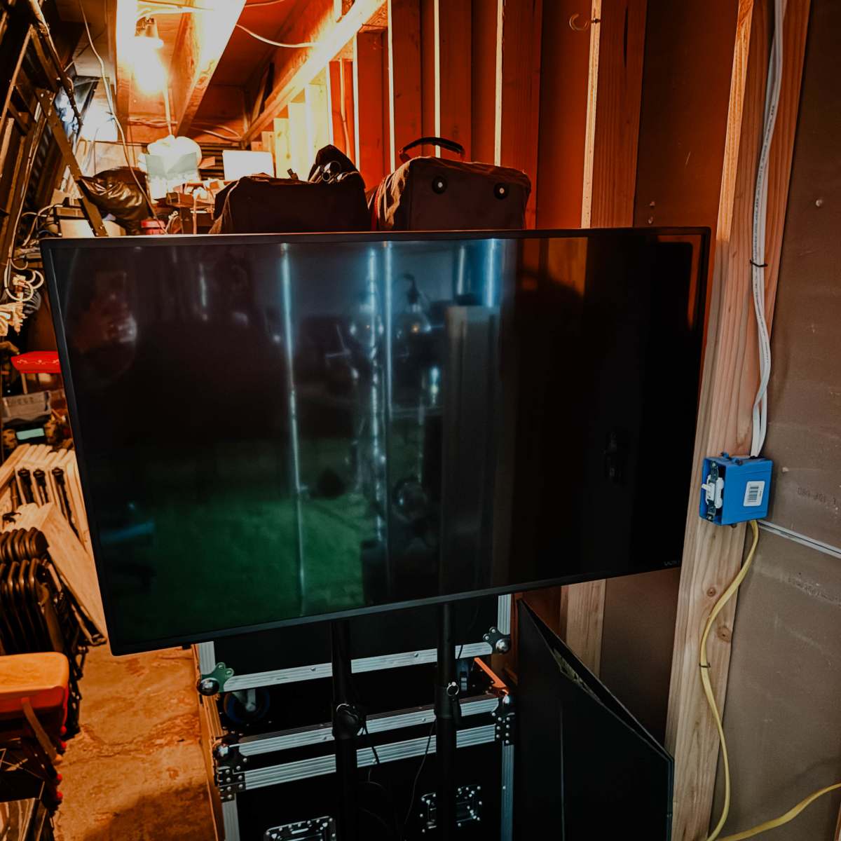 A flat-screen television is mounted on a stand in a cluttered storage area filled with equipment and bags.