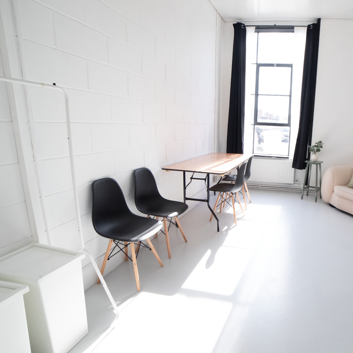 A bright, minimalistic room featuring a sofa, a wooden table with chairs, a plant, and a window.